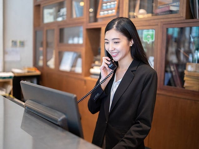 A woman in a black suit is standing at a reception desk, speaking on a landline phone and smiling. She is in front of a computer monitor, with shelves lined with books and decor visible in the background.