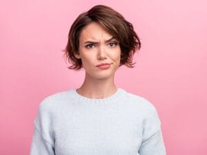A person with short brown hair wearing a light gray sweater stands against a pink background, displaying a quizzical expression with one eyebrow raised.