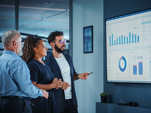 Three people in an office setting analyze graphs and data displayed on a large screen. A man points to the screen while discussing the information. The atmosphere is collaborative and professional.