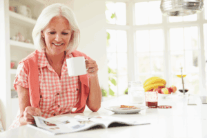 Older woman sitting at a table sipping from a mug while reading a magazine and smiling