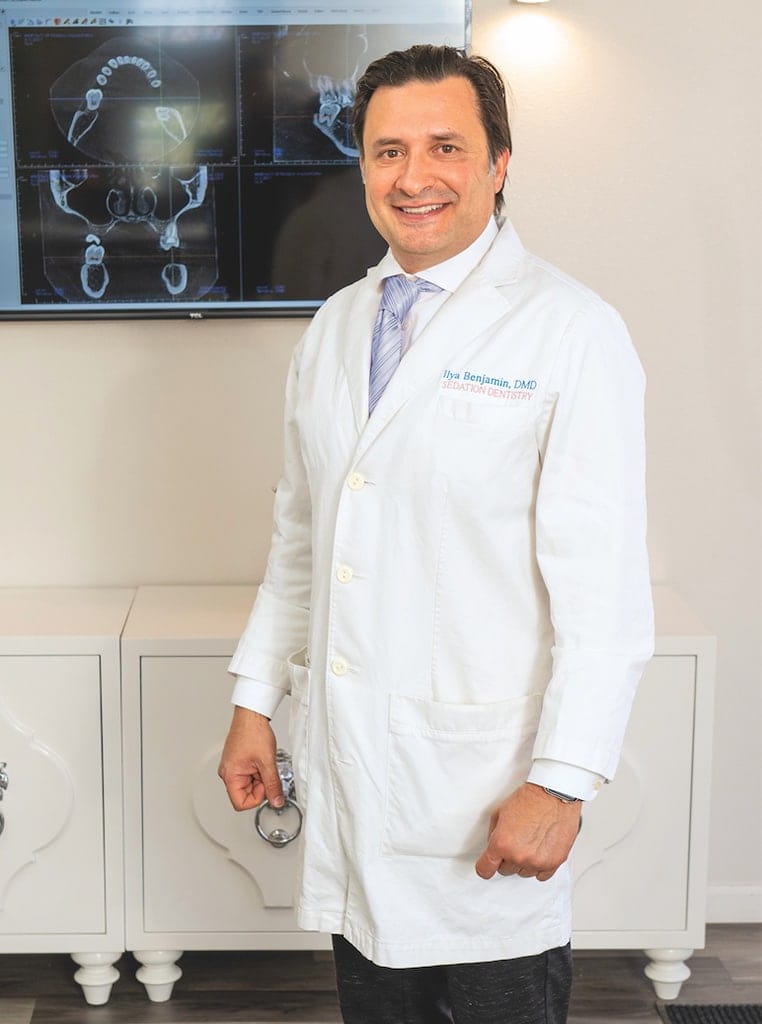 A dentist standing and smiling, wearing a white coat with Dr. Ben embroidered. Behind him is a screen displaying dental X-rays, and a cabinet with a polished surface. The setting appears to be a dental office.