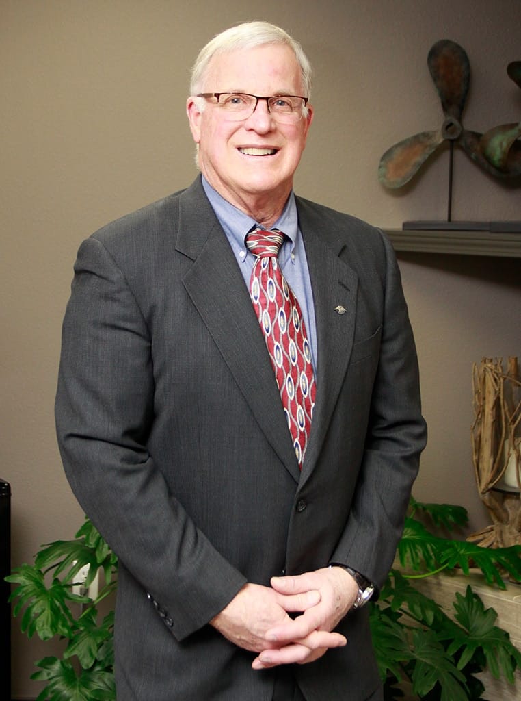 Rick Raney, a man with glasses, gray hair, and a smile, stands confidently in an office. He is wearing a dark suit, blue shirt, and patterned tie. Behind him, theres a plant and a decorative object on a shelf, adding charm to the space.